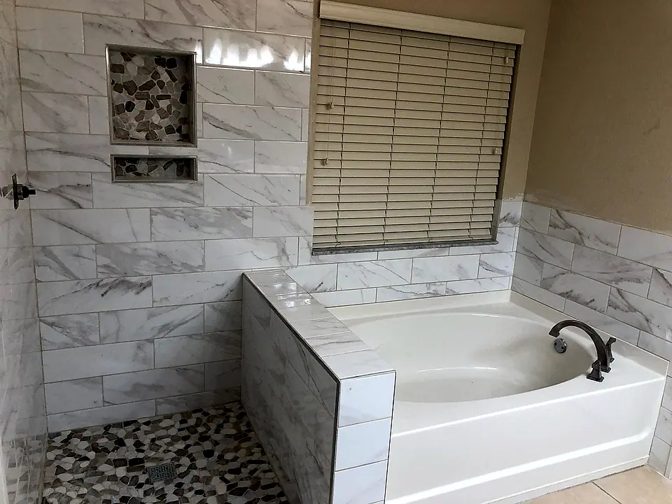 The same primary bath finished with white marble-look tile, a pebble shower floor and a tiled tub deck